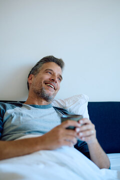 Man smiling, drinking coffee while relaxing in bed during morning, feeling happy and content with lifestyle