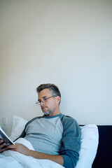 Fototapeta premium Mature man in pajamas relaxing in bed reading a magazine, enjoying a peaceful early morning of quiet leisure and self-care