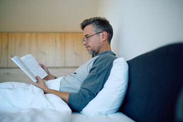Man relaxing in bed, wearing glasses, reading a paper book. Enjoying quiet morning leisure time at home