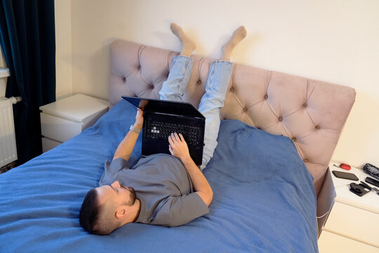 A guy is lying on his bed with his legs up, using a laptop for his online job. The bedroom is neat, and he seems comfortable while working from home and taking a break