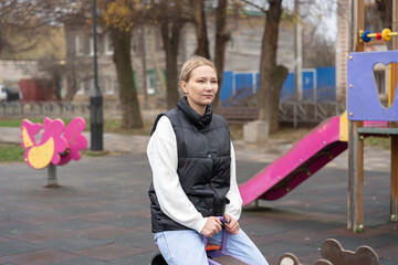 A young woman sits on a spring rider at an empty children's playground, quietly remembering her childhood with a nostalgic, reflective mood amid autumn trees and muted urban surroundings