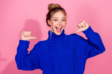 Young woman in blue knit sweater smiles and gives thumbs up against pink background