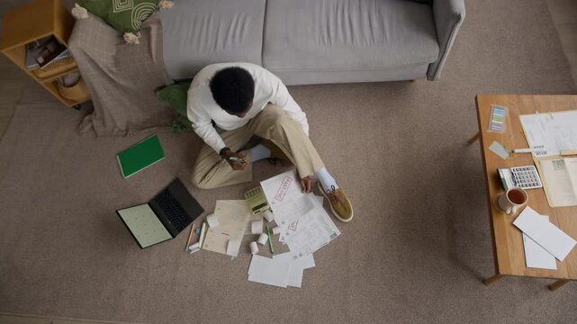 Top down view shot of African American man organizing receipts and unpaid bills on floor, calculating monthly expenses with calculator at home, showing financial concern