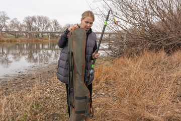Fototapeta premium Female angler packing a fishing rod into a carry case by a calm river shore in autumn