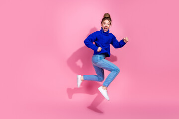 Jumping with joy a young woman in a blue sweater and jeans against a pink background