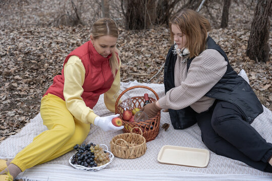 Two young women enjoy an autumn picnic in a quiet forest, sitting on a light blanket among fallen leaves, sharing apples, grapes and wine from a wicker picnic basket while wearing cozy vests - Powered by Adobe