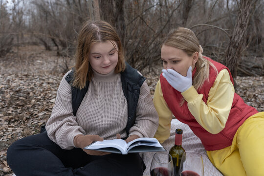 Two women reading book outdoors on a picnic blanket, expressing astonishment at a plot twist while wrapped in warm sweaters and gloves near bare trees