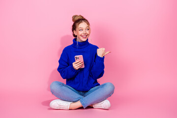 Young woman sits cross legged on pink background smiling as she uses a pink phone dressed in blue...