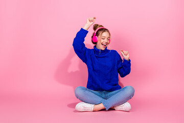 Young woman in blue sweater wearing pink headphones dances happily against pink backdrop