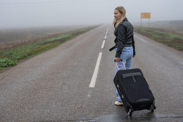 Solitary traveler on a misty rural road dragging a suitcase in a leather jacket and blue jeans, emotional moment of departure and loneliness