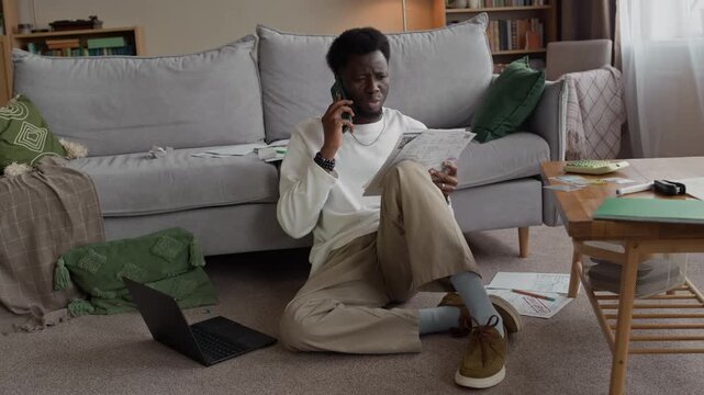 Frustrated Black man at home arguing on phone while reviewing bills and finances with stressed expression, managing household expenses and overdue payments