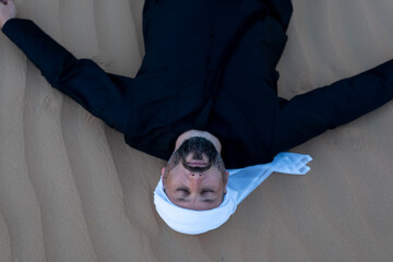Relaxing male tourist wearing traditional arab clothing, lying on desert sand dunes. High quality photo