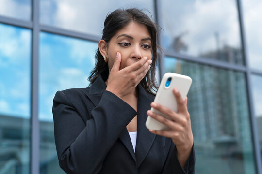 Woman in business attire gasps at news on phone outside modern building during daytime