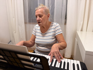 An elderly woman playing a keyboard in a cozy room, focused on the music sheet, wearing a striped shirt, with light-colored hair tied back