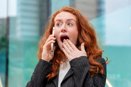 Excited businesswoman reacts with surprise while talking on the phone outside modern building in city