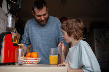 Happy boy tasting freshly squeezed juice. Father and son preparing fresh orange juice using modern screw electric juicer in kitchen. Healthy morning routine, family life, housework help.