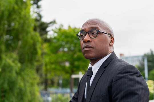 Man in a black suit stands confidently outdoors against a backdrop of lush greenery in springtime - Powered by Adobe