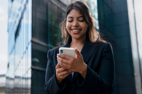 Business professional smiles while using smartphone outside modern office building during daylight