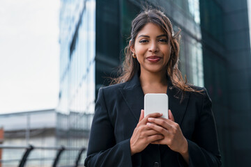Confident woman holding smartphone in city near modern buildings during sunny day