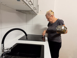 An elderly woman in a kitchen, cleaning a black glass cooktop while holding a bowl