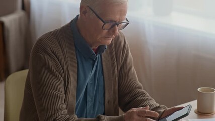Tilt up of elderly Caucasian man checking bills and managing finances using smartphone at wooden desk in home setting - Powered by Adobe