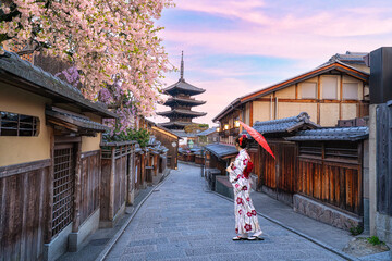 A young Japanese woman in a traditional kimono stands beside the Yasaka Pagoda in Kyoto, Japan, amidst blooming cherry blossoms. © munduuk