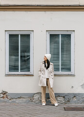 Woman in a stylish beige trench coat and white beanie stands against a textured wall with windows, showcasing a modern urban fashion look with copy space