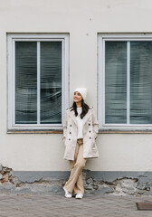 Young woman dressed in stylish winter attire, standing confidently against a textured wall with windows, showcasing a modern urban fashion aesthetic and casual elegance