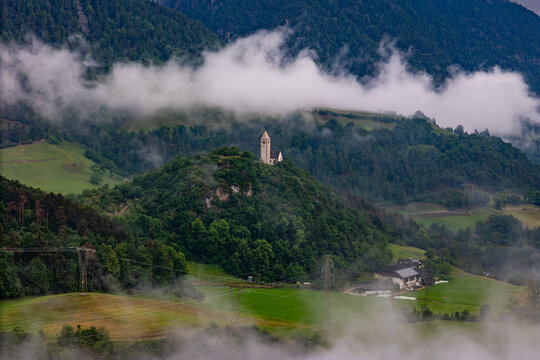Church of Saint Verena on a wooded hill with low cloud and valley mist, aerial view. Dolomites, South Tyrol, Italy