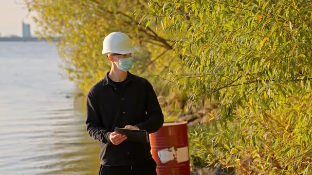 Scientist In PPE Suit, Hard Hat, And Face Mask Writing Down His Evaluation After Inspecting The Trees On The Banks Of River. - wide shot