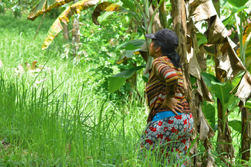 A mother stands in tall grass near banana trunks, hand on hip, looking intently to the side in the...