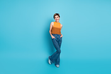 Stylish young woman in orange top and blue jeans posing against bright blue studio background