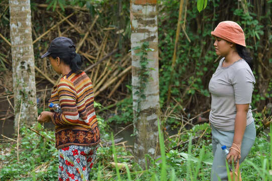 A mother with a water bottle walks past tree trunks near a river, followed by her teenage daughter carrying another bottle in the agroforestry field. - Powered by Adobe