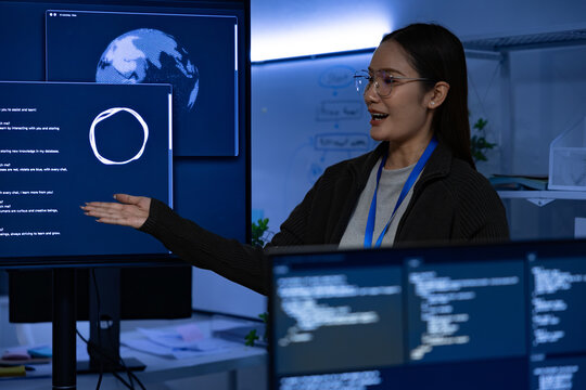 Young woman presenter pointing at large monitor with globe and diagram, smiling and explaining technology concept in modern office