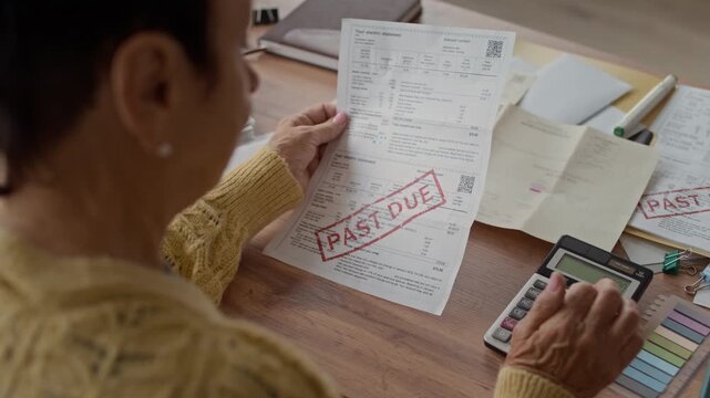Over the shoulder shot of elderly woman sitting at home desk holding overdue bill while using calculator to manage household finances