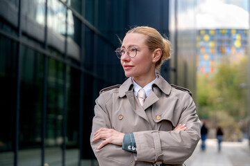 Confident woman in a stylish trench coat stands in an urban area with modern architecture © Iryna