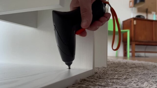 Hand with cordless electric screwdriver that screws for a clean and secure fit, tightening the screws of a piece of furniture at home