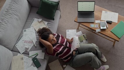 Top down view shot of worried Biracial woman sitting on floor at home surrounded by stacks of overdue bills using laptop to manage finances and plan payments - Powered by Adobe