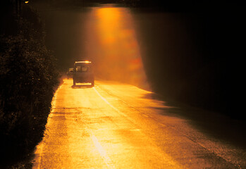 Car driving along a rural road illuminated by intense golden sunlight creating a dramatic warm and...