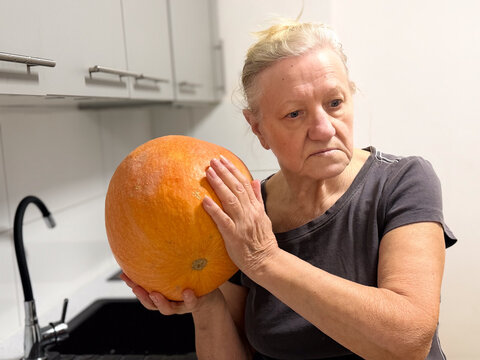 A woman holding a large pumpkin in a kitchen setting, looking contemplative, with a neutral expression - Powered by Adobe