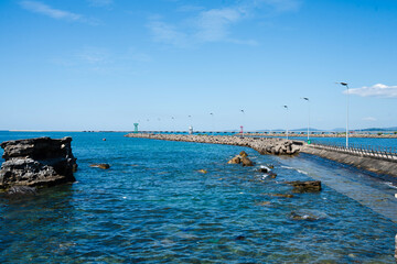Fototapeta premium A clear blue seaside scene with rocky shores, calm waves, and a long breakwater stretching into the distance under a bright sky.