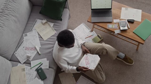 Top down view shot of depressed Black man sitting on floor among stacks of overdue bills, leaning his head back on sofa, showing stress, worry, and emotional burden of debt