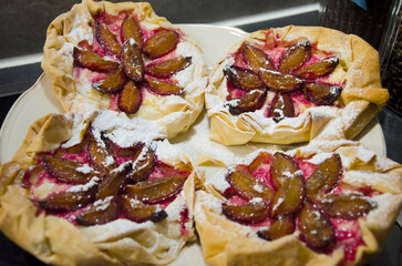 Close-up of freshly baked homemade plum tarts made with puff pastry, topped with sliced plums arranged in a floral pattern and dusted with powdered sugar.