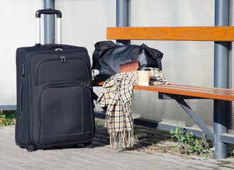 Black suitcase, travel bag, checked scarf, and takeaway coffee cup placed on a wooden bench at a bus stop.