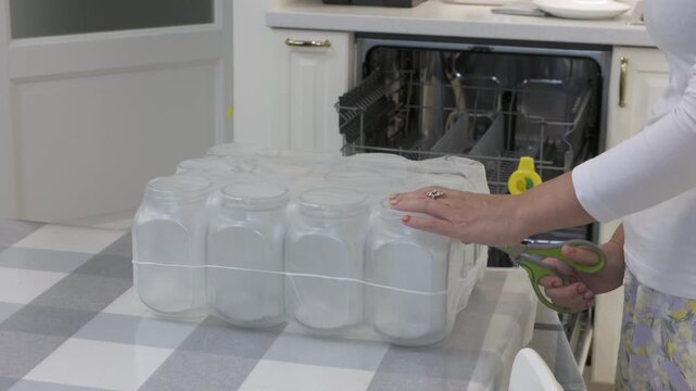 A woman uses scissors to cut the plastic packaging of new glass jars. She stands at a table with a checkered cloth in a modern kitchen with an open dishwasher.