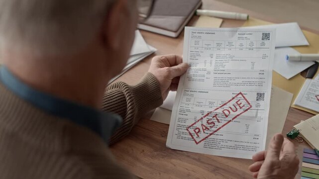 Over the shoulder shot of older man sitting at table reviewing household bill stamped with red past due while organizing personal finances in home setting