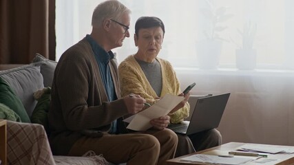 Medium shot of senior Caucasian couple reviewing bills and planning budget together on laptop while sitting on sofa in warm and comfortable home environment - Powered by Adobe
