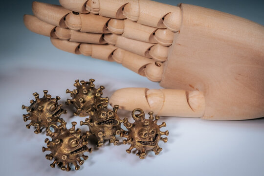 The hand of a wooden mannequin hangs above several bronze virus-shaped figurines with faces on a light surface. - Powered by Adobe