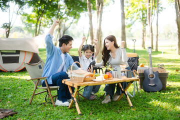 Joyful Asian family camping in a sunny garden, bonding over BBQ, tech, and laughter. Parents and daughter enjoy outdoor