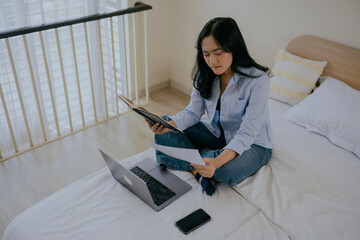 Serious asian woman sit on bed, check household bills, makes online payment using e-bank laptop...